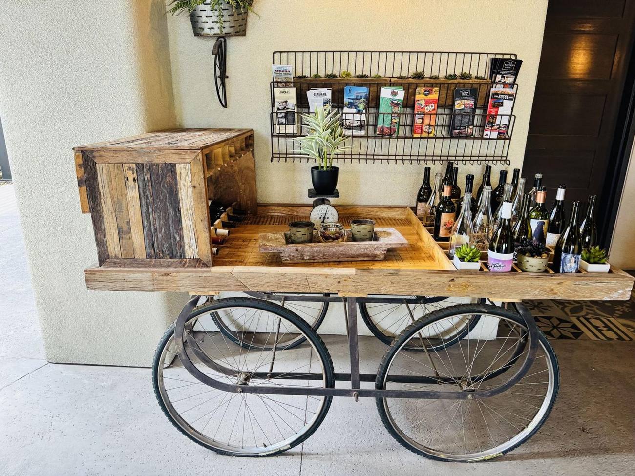 A rolling wooden cart with wine bottles, small plants, and gift items outside a shop in Old Town Cottonwood