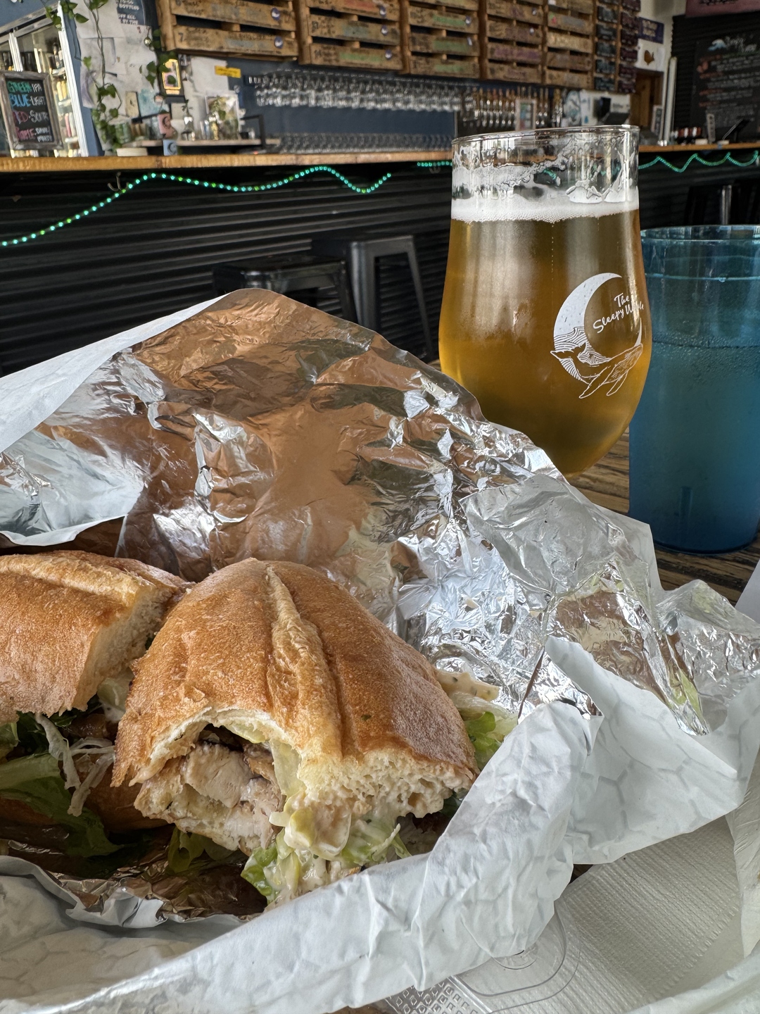 A pint glass with the Sleepy Whale logo, a foil-wrapped sandwich from Best Pizza and Brew, and a blue plastic cup filled with water for Dexter — all together on the taproom table.