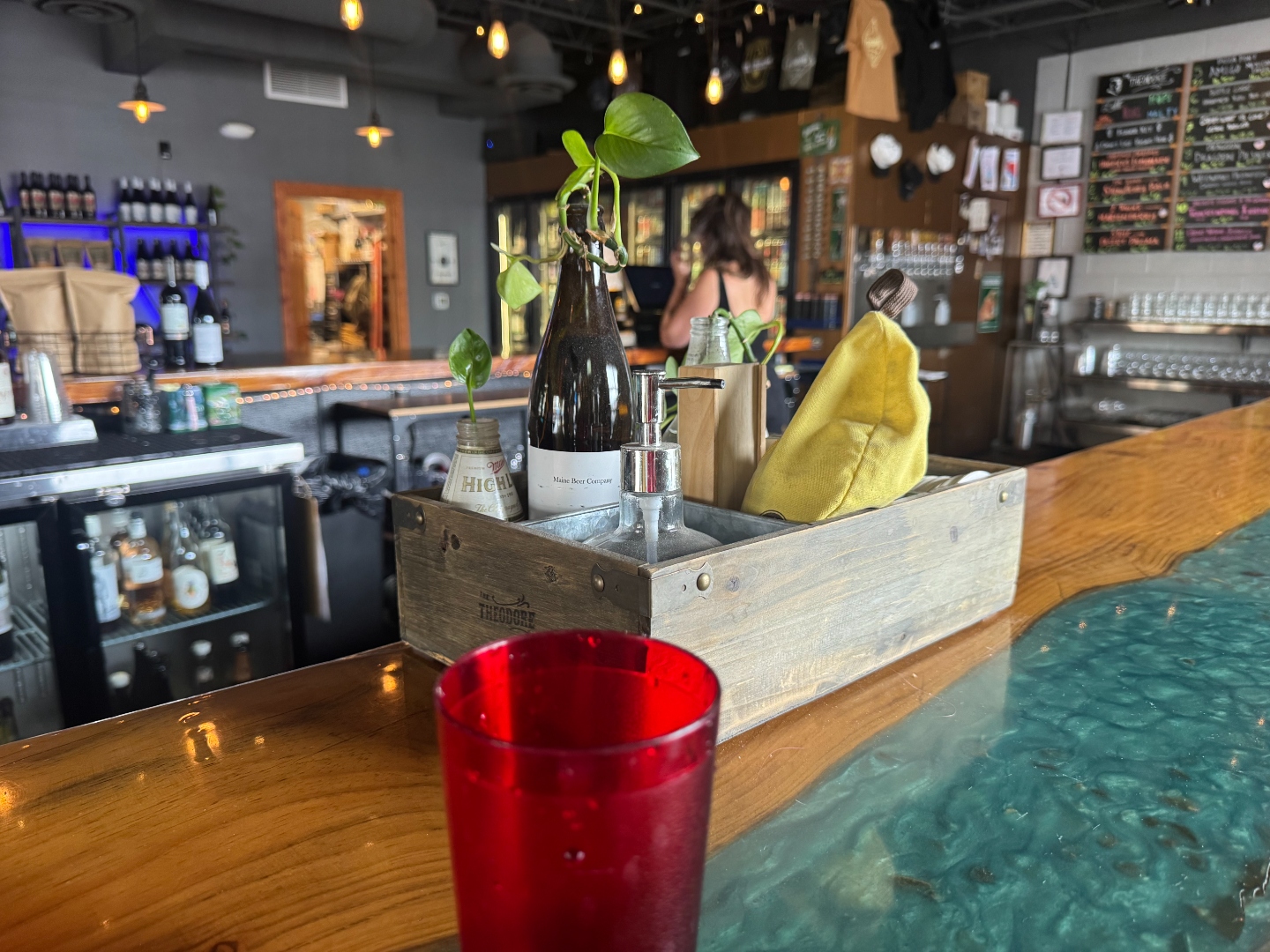 A wooden Theodore-branded bar caddy on the turquoise resin bar, holding a Maine Beer Company bottle, a soap pump, and a yellow cloth. A red water tumbler sits in the foreground.