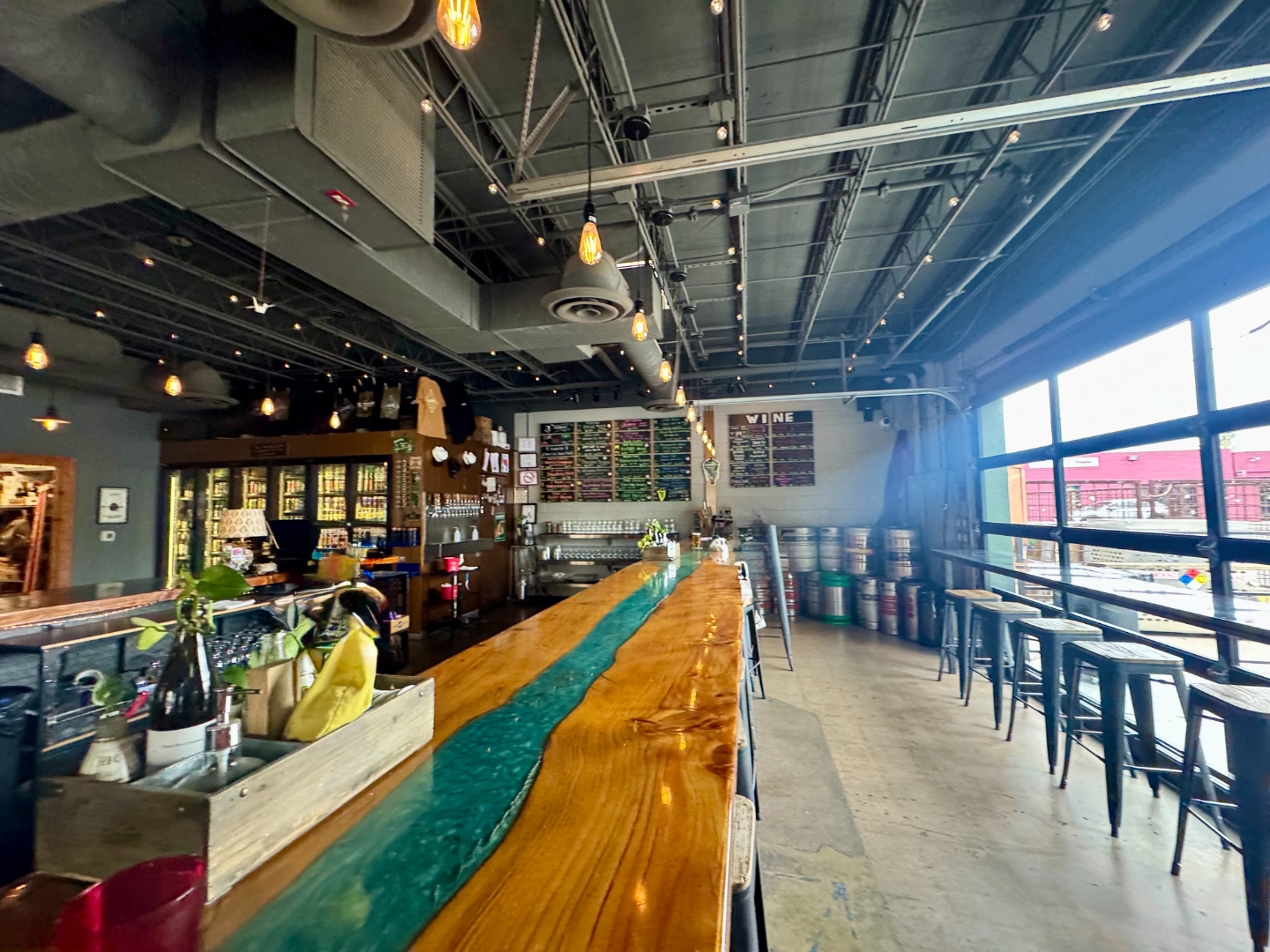 The Theodore taproom interior, downtown Phoenix. A long live-edge wooden bar with a turquoise resin river running down the middle, industrial ceilings with pendant Edison bulbs, and a wall of chalkboard beer and wine lists above the taps.