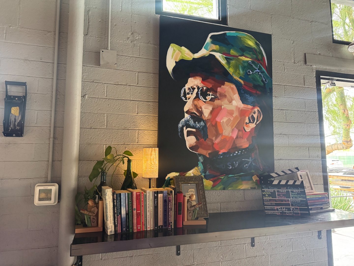 A large painted portrait of a bearded man in a green cap, mounted above a shelf of books and a Teddy Film Club clapperboard at The Theodore.